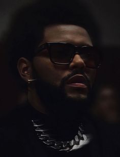 a man wearing sunglasses and a chain around his neck looks off into the distance with dark lighting behind him