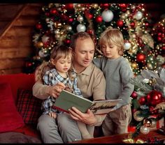 a man sitting on a couch with two children reading a book in front of a christmas tree