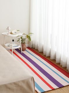 a room with a bed, table and colorful striped rug on the floor in front of a window