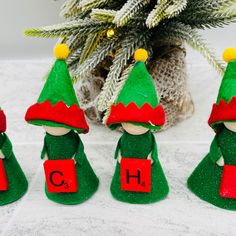 three green elfs with red hats and the letters ch on their backs are next to a potted plant