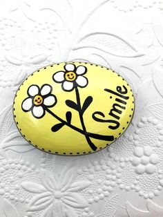 a painted rock that says smile on it with flowers and leaves in the center, sitting on a white surface