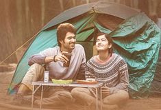 a man and woman sitting on the ground in front of a tent with a cup of coffee