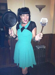 a woman in a green dress holding two cooking utensils and a frying pan