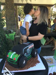 a woman kissing a small child on the cheek in front of a cake with a monster truck on it