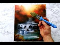 someone is holding a paintbrush in front of a painting with waterfall and sun on it