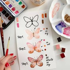 a woman's hand holding a paintbrush next to some watercolors