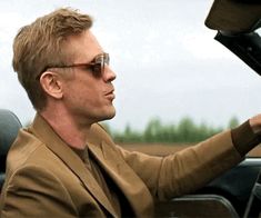 a man wearing glasses driving a car with his hand on the steering wheel and looking out the window