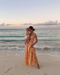 a woman holding a baby standing on top of a sandy beach next to the ocean