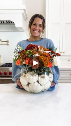 a woman holding a vase filled with flowers and pumpkins on top of a kitchen counter