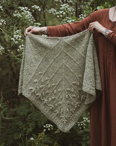 a woman is holding up a crocheted shawl in front of some flowers