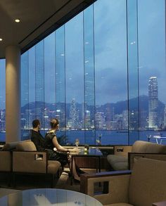 two people sitting on couches in front of large windows looking out at the city