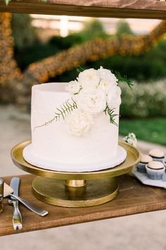 a wedding cake with white flowers and greenery sits on a gold platter next to cupcakes