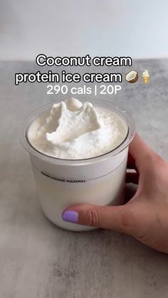 a hand holding a container of cream on top of a white counter next to a cup with whipped cream in it