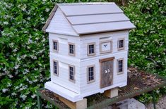 a white doll house sitting on top of a wooden platform in front of some bushes