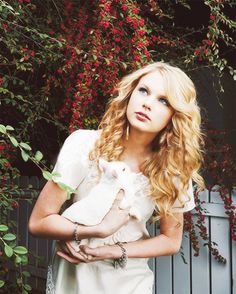 a woman holding a small white dog in her arms next to a bush with red flowers