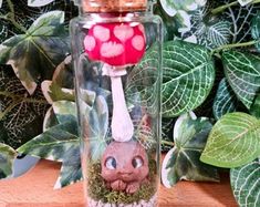 a glass jar with a red mushroom on the top and green leaves around it, next to a plant