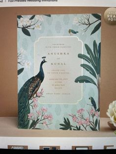 a wedding card with a peacock on it and flowers in front of the card is shown