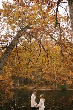 a man and woman standing next to each other in front of a lake surrounded by trees