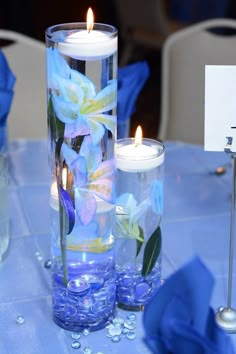 two candles are sitting in glass vases with blue flowers and water on the table