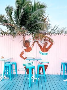 two people sitting at blue tables with palm trees in front of pink wall and wooden flooring