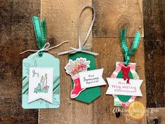 three tags are hanging on a wooden surface with ribbons and ribbon around them, one has a christmas stocking ornament