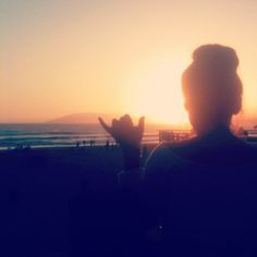 a person sitting in front of the ocean with their hand up to the sun setting