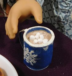 a doll is holding a cup of hot chocolate with marshmallows in it