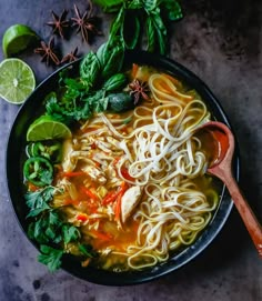 a bowl of chicken noodle soup on a table with limes and an orange slice