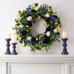 a wreath with blue and white flowers sitting on top of a mantle next to candles