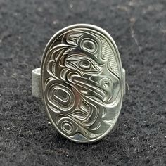 On offer is a West Coast Namgis First Nation custom made to order hand carved sterling silver 'Kulus' plaque ring. by World renowned master carver Joe R. Wilson with his depiction of Kulus in the style of the Namgis, Kwakiutl First Nation. Initialed by Joe. Fine example of Indigenous Art from the Northwest Coast First Nations of Canada. PLEASE NOTE THAT THIS IS A CUSTOM RECREATED HAND CARVED ITEM THAT WILL HAVE SLIGHT, SUBTLE DIFFERENCES THAT ARE VERY HARD TO DISTINGUISH WHEN COMPLETED FROM THE Symbolic Carved White Gold Jewelry, Symbolic Carved Sterling Silver Rings, Symbolic Carved Engraved Ring As Gift, Symbolic Engraved Carved Ring As Gift, Symbolic Engraved Carved Ring For Gift, Artisan Oval Engraved Ring, Oval Sterling Silver Engraved Ring, Ceremonial Silver Carved Engraved Ring, Ceremonial Sterling Silver Engraved Carved Ring