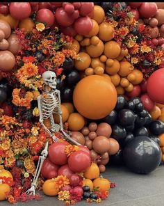 a skeleton sitting on top of a pile of balloons and flowers in front of an orange wall