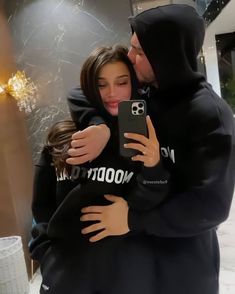 a man in a hoodie hugging a woman who is holding her phone up to her face