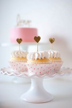 two cupcakes with hearts on top sitting on a cake stand in front of a pink and white cake