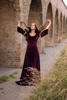 a woman in a long purple dress is standing on a brick walkway and has her hands behind her head