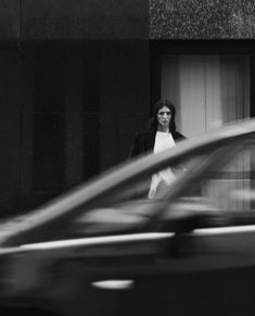 a black and white photo of a woman standing in front of a building next to a car