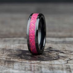 a pink and black wedding ring with red glitter inlaying the center, on top of a piece of wood