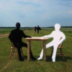 two people sitting at a table in the middle of a field with one person standing