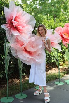 a woman in white dress standing next to large pink flowers with green stems on each side