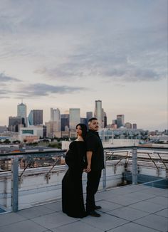 a man and woman standing next to each other on top of a roof looking at the city