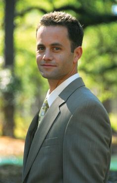 a man wearing a suit and tie standing in front of some trees with his head turned to the side