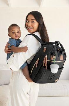 a woman holding a baby in her back pack while standing next to a white couch