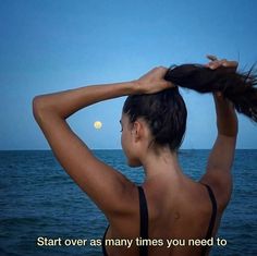 a woman standing in front of the ocean holding her hair up to her head and looking at the moon