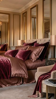 a large bed with maroon sheets and pillows in a bedroom next to a mirror on the wall