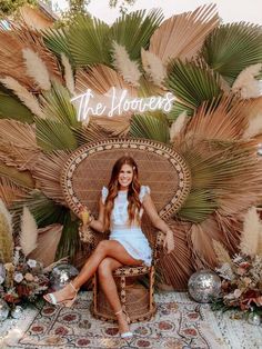 a woman sitting on top of a wicker chair in front of a wall with palm leaves