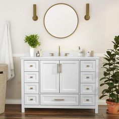 a white bathroom vanity with gold handles and round mirror above it, next to a potted plant