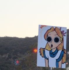 there is a sign with a painting of a woman wearing sunglasses on the side of a hill