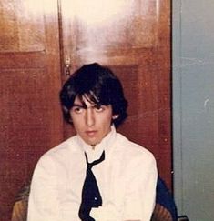 a young man wearing a white shirt and black tie sitting in front of a wooden door