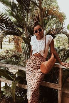 a woman sitting on top of a wooden bench next to palm trees and holding a purse