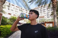 a young man drinking from a can while standing in front of a palm tree and building