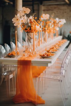 a long table with clear chairs and orange flowers in vases on the top tables